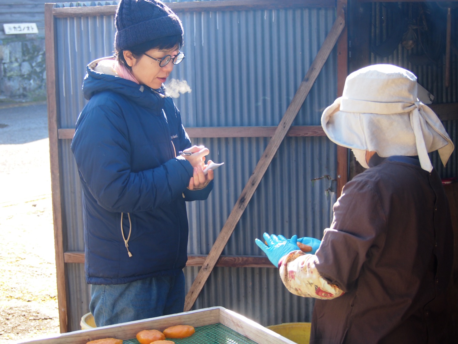 干し芋作業をしている恵子さんに店主の小清水が取材をしています